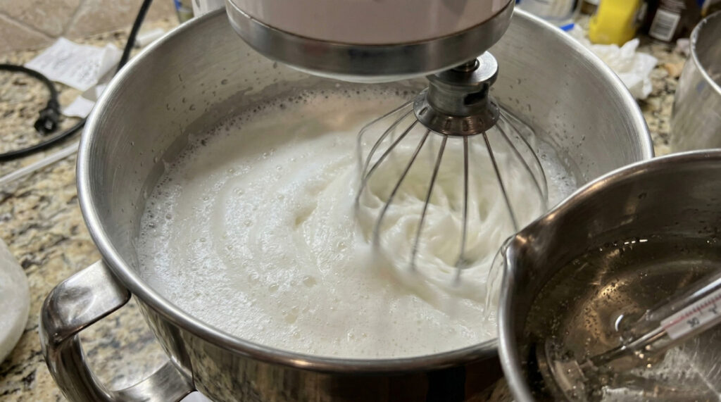 Stand mixer whisking egg whites to soft, cloudy peaks in a stainless steel bowl.